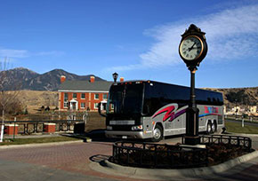 Big charter bus parked next to clock post with mountains in background.
